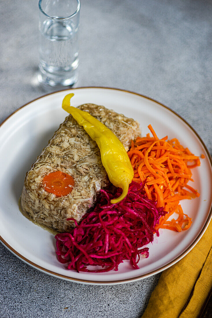 Top view of chicken aspic garnished with a carrot slice, accompanied by spicy shredded carrot and pickled beetroot cabbage salad, presented with a clear glass of vodka on a white plate