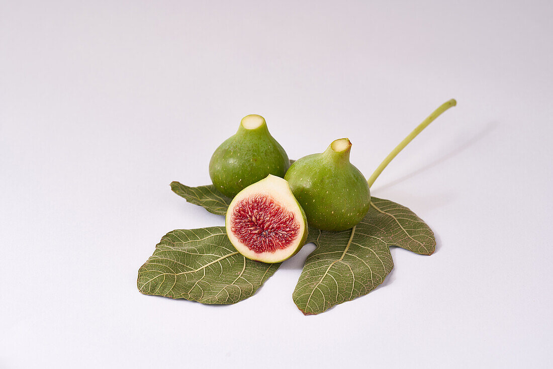 A composition of ripe green figs, one sliced in half, with a detailed leaf on a clean white surface.