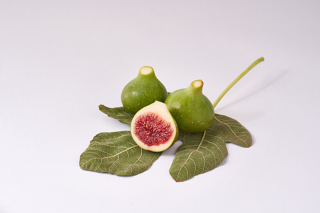 Eine Komposition aus reifen grünen Feigen, eine in Scheiben geschnitten, mit einem detaillierten Blatt auf einer sauberen weißen Oberfläche