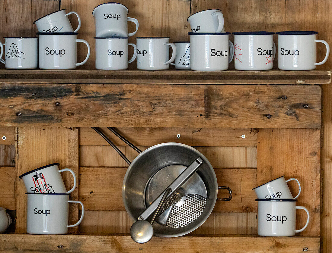 Weiße Tassen mit dem Schriftzug "Soup" stehen ordentlich auf einem Holzregal, begleitet von einem Sieb und einer Schöpfkelle, und vermitteln eine rustikale und einladende Küchenatmosphäre