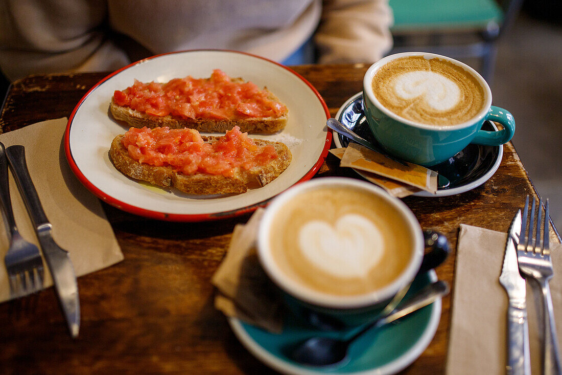 Köstliche, appetitliche Toasts mit Tomaten auf einem Teller neben Tassen mit aromatischem Cappuccino auf einem Holztisch in einer Cafeteria