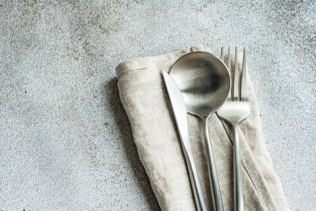 Besteckset auf grauer Oberfläche vor rauem, strukturiertem Hintergrund mit Handtuchserviette und trockenem Löffel in heller Küche
