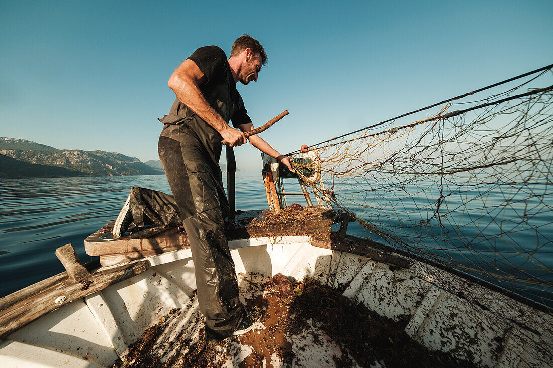 Von unten fokussierter bärtiger männlicher Fischer in Uniform, der mit einem Netz auf Fischfang geht, während er auf einem Schoner in Soller nahe der Baleareninsel Mallorca arbeitet