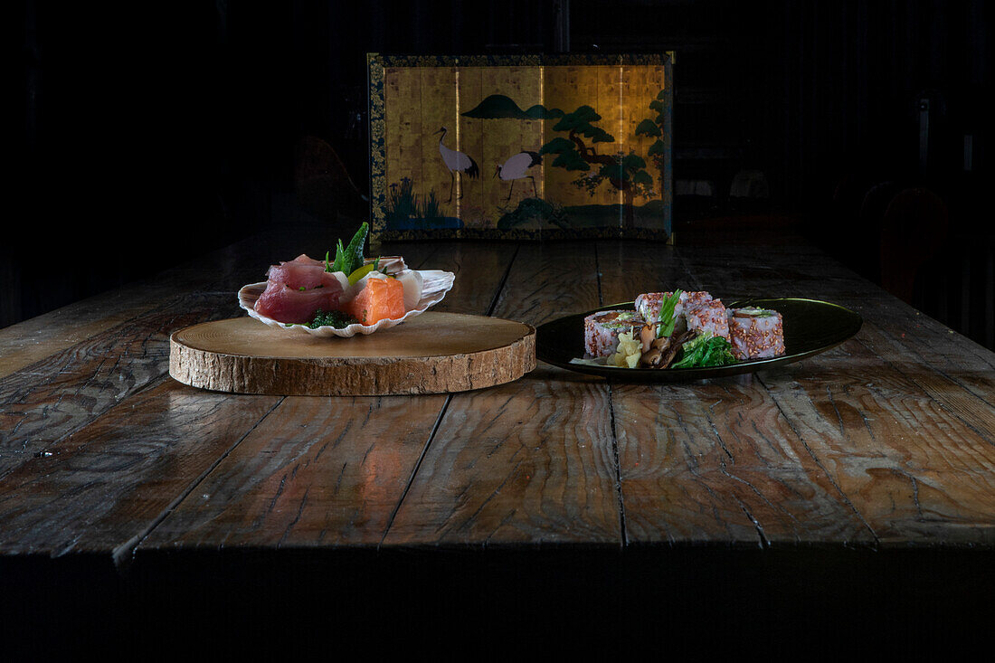 Appetizing sashimi of raw tuna and salmon in white plate on wood slice while sushi rolls made with rice avocado fish in black plate and served on table against dark background