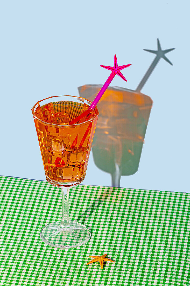 Ein eleganter orangefarbener Cocktail mit einem rosafarbenen Sternrührer steht auf einem grün karierten Tuch vor einem blassblauen Hintergrund und wirft einen verspielten Schatten