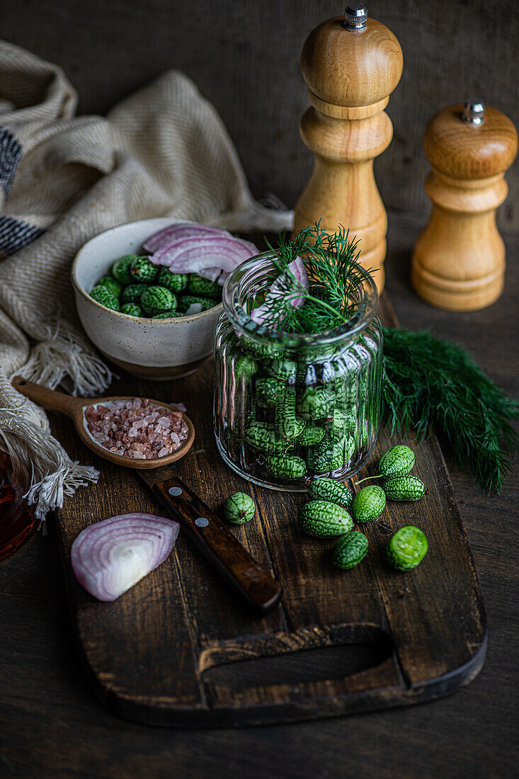 High angle of ingredients for preparing cucamelon fermentation placed in jar placed on wooden tray near napkin and salt and pepper shakers against dark background