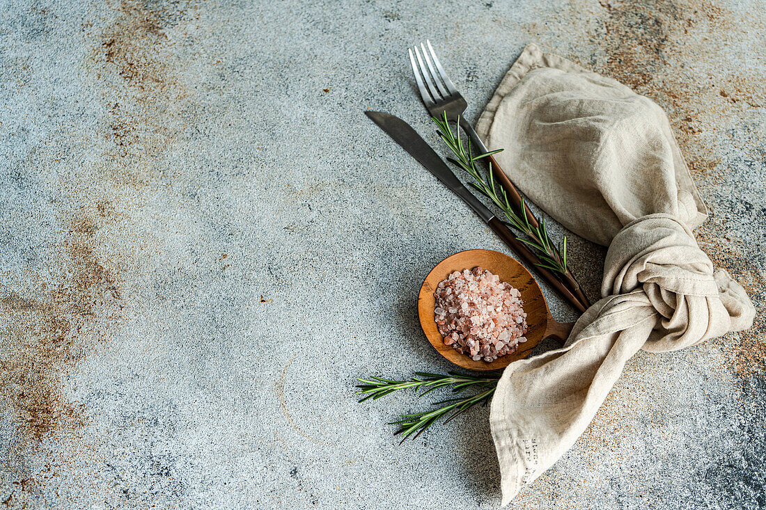 From above wooden spoon with pink Himalayan salt, surrounded by rosemary sprigs and a folded linen napkin alongside a knife and fork, all arranged neatly on a textured concrete surface