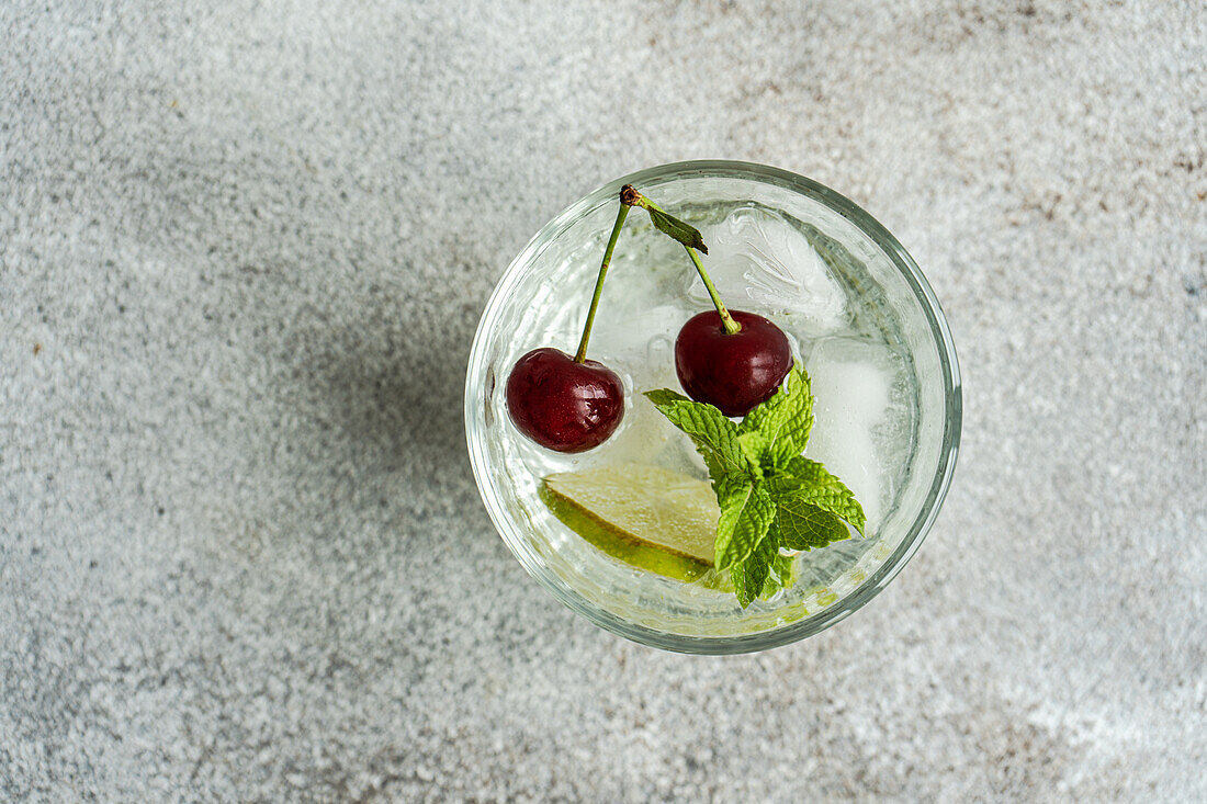 From above transparent glass of refreshing cold drink with lime and cherry and mint leaves on gray surface in summer day against blurred background