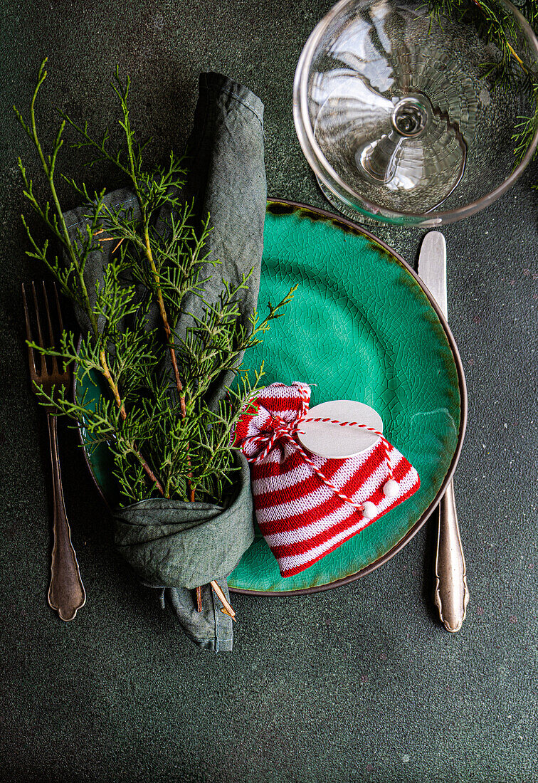 Draufsicht auf eine kleine Weihnachtstüte und in ein Taschentuch gewickelte Tannenzweige auf einem Teller, der auf einem grünen Tisch neben Glas und Besteck steht