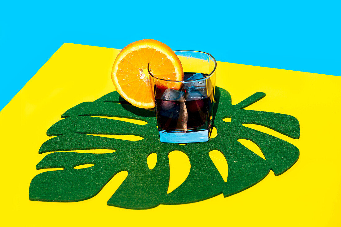 Kaltes Getränk mit Orangenscheibe im Glas auf Blattdekor über gelbem Hintergrund vor blauem Hintergrund im Studio