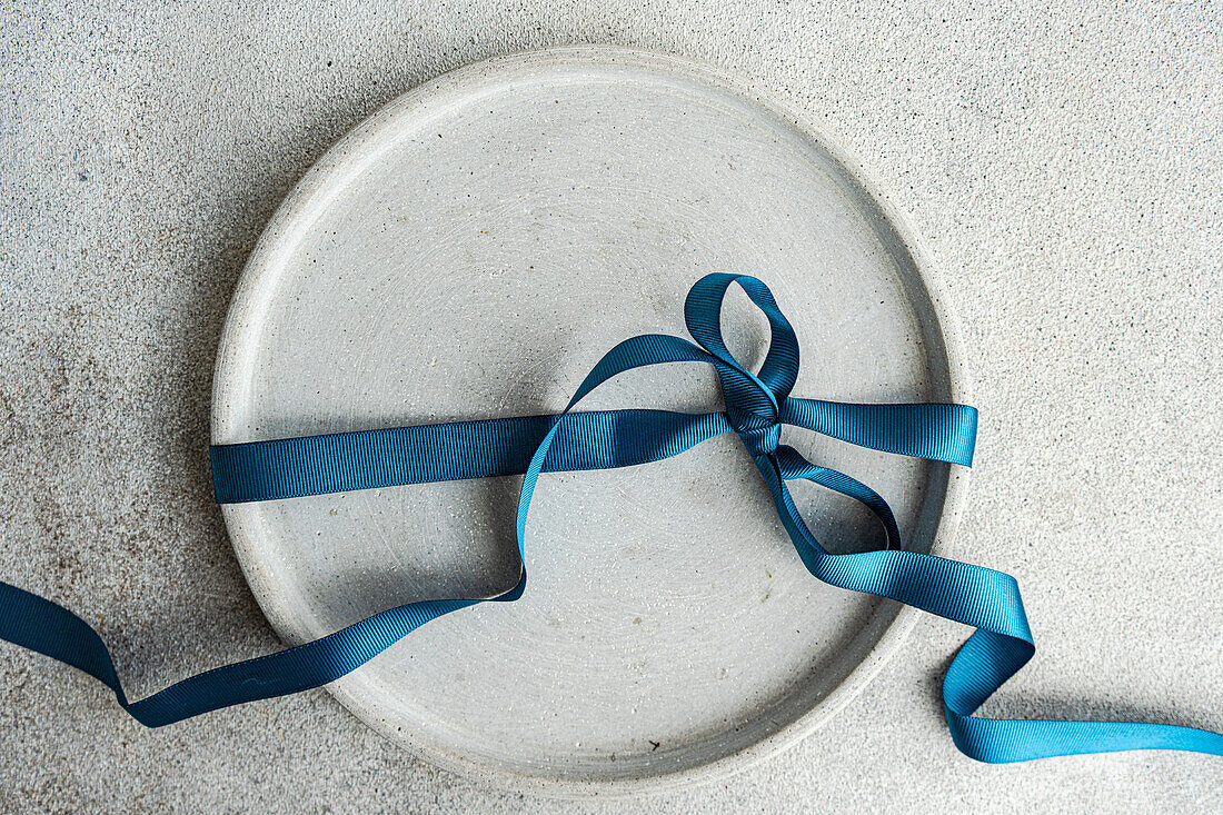 Draufsicht auf einen minimalistisch und doch stilvoll gedeckten Ostertisch mit einem grauen Teller, der mit einem blauen Band gebunden ist, auf einer grauen Fläche