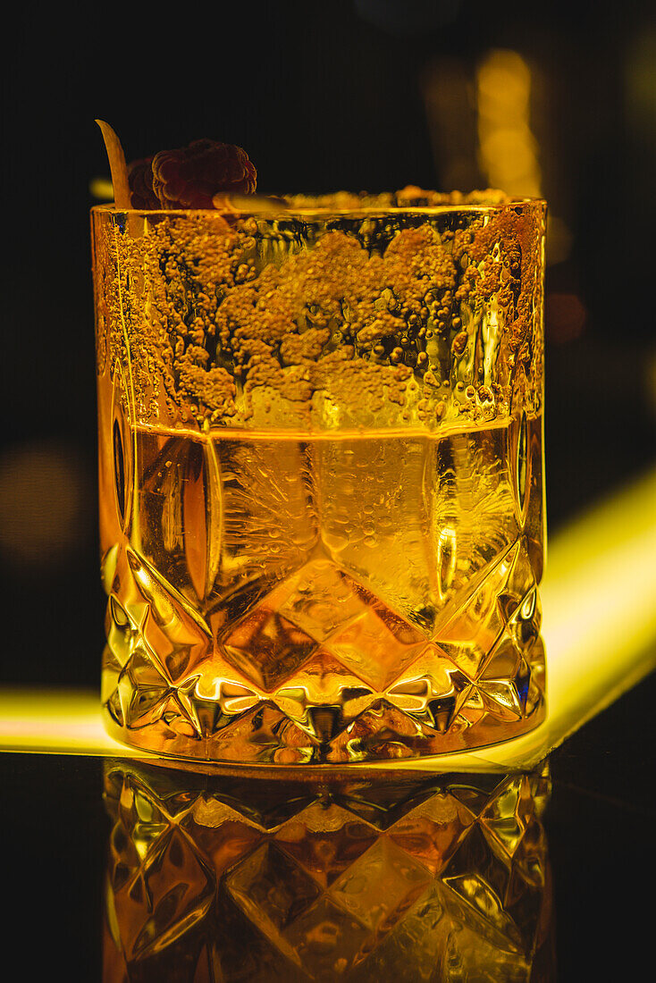 Ein mit Whiskey gefülltes Glas steht auf einem reflektierenden Tisch mit gelber Beleuchtung in einer dunklen Bar