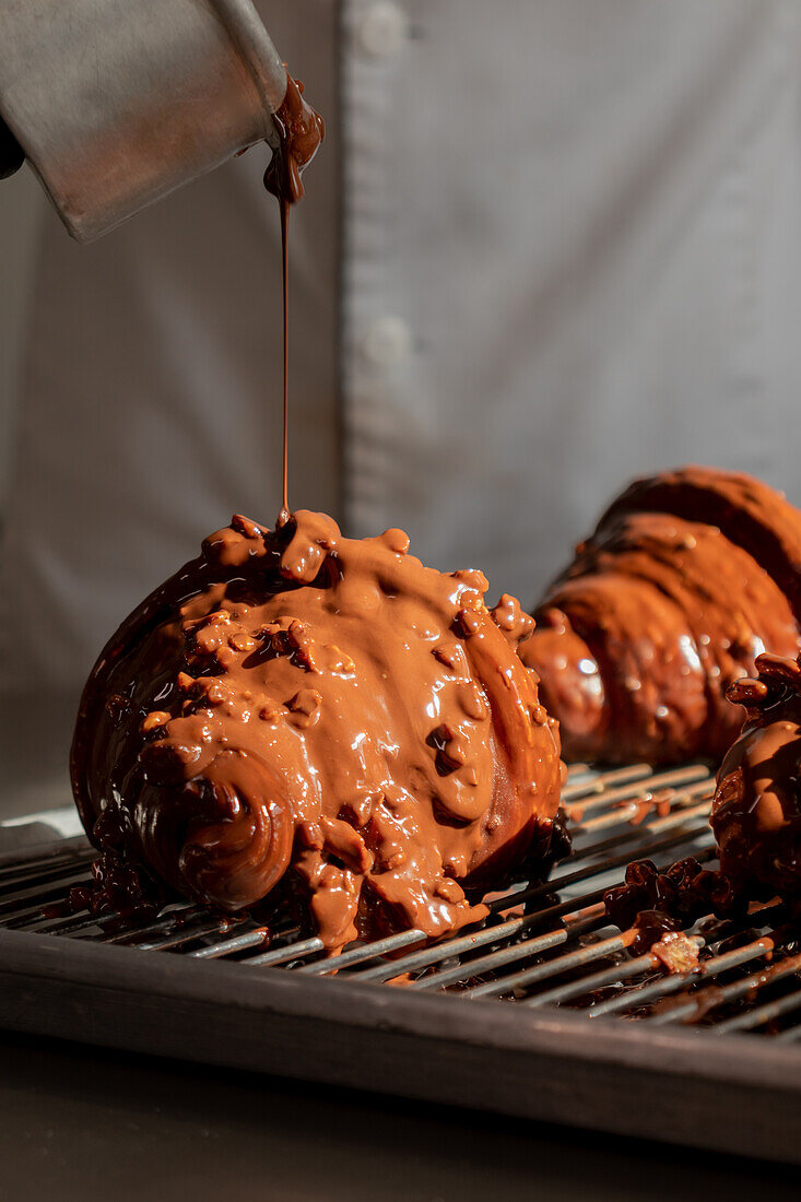 Unrecognizable person pouring chocolate glaze on appetizing croissants placed on metal tray while preparing pastries in factory