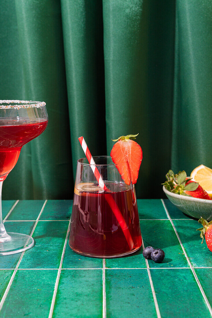 Gläser mit erfrischenden roten alkoholischen Cocktails mit Erdbeeren und Strohhalm, serviert auf einem Tablett mit Beeren und Zitronenscheibe auf dem Tisch