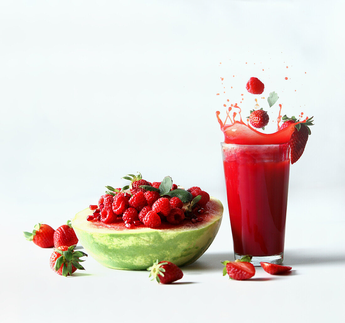 Spritzendes Getränk mit Wassermelone, serviert mit roten Sommerfrüchten, Beeren und rotem Saft: Erdbeeren und Himbeeren fallen in Glas auf weißem Hintergrund. Gesunder erfrischender Sommersnack Vorderansicht