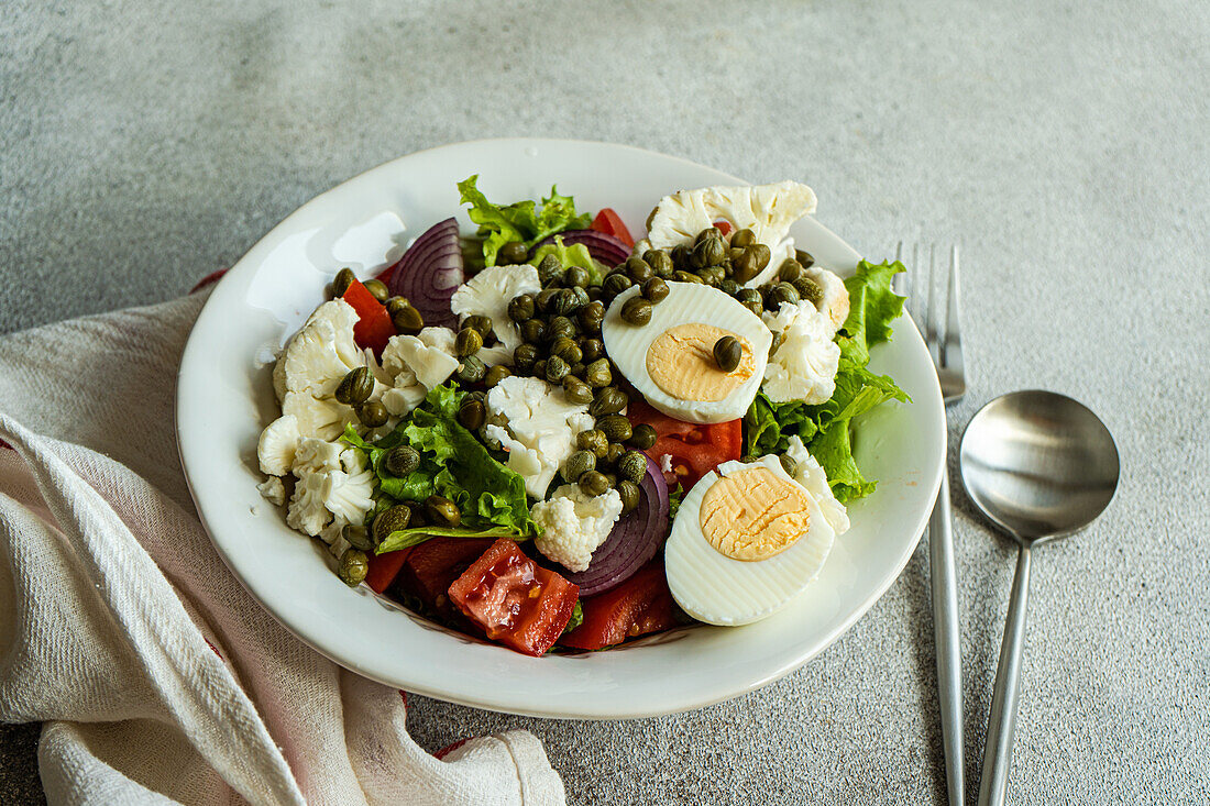 Hoher Winkel eines Tellers mit Keto-Salat mit Salatblättern, gekochten Eiern, Kapern, Tomaten, roten Zwiebeln und Blumenkohlgemüse auf dem Tisch zwischen Serviette und Besteck