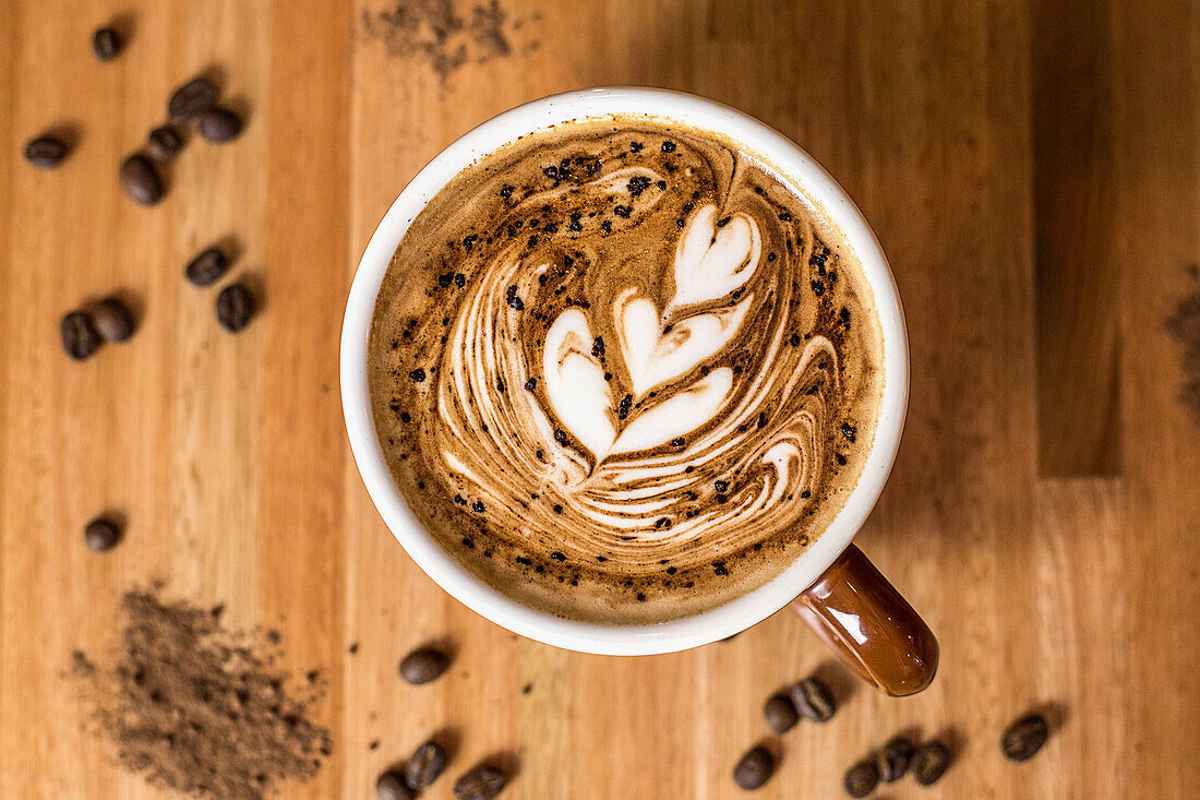 Top view of ceramic mug with fresh aromatic creamy latte coffee placed on wooden dinning table with dry coffee seeds in daylight