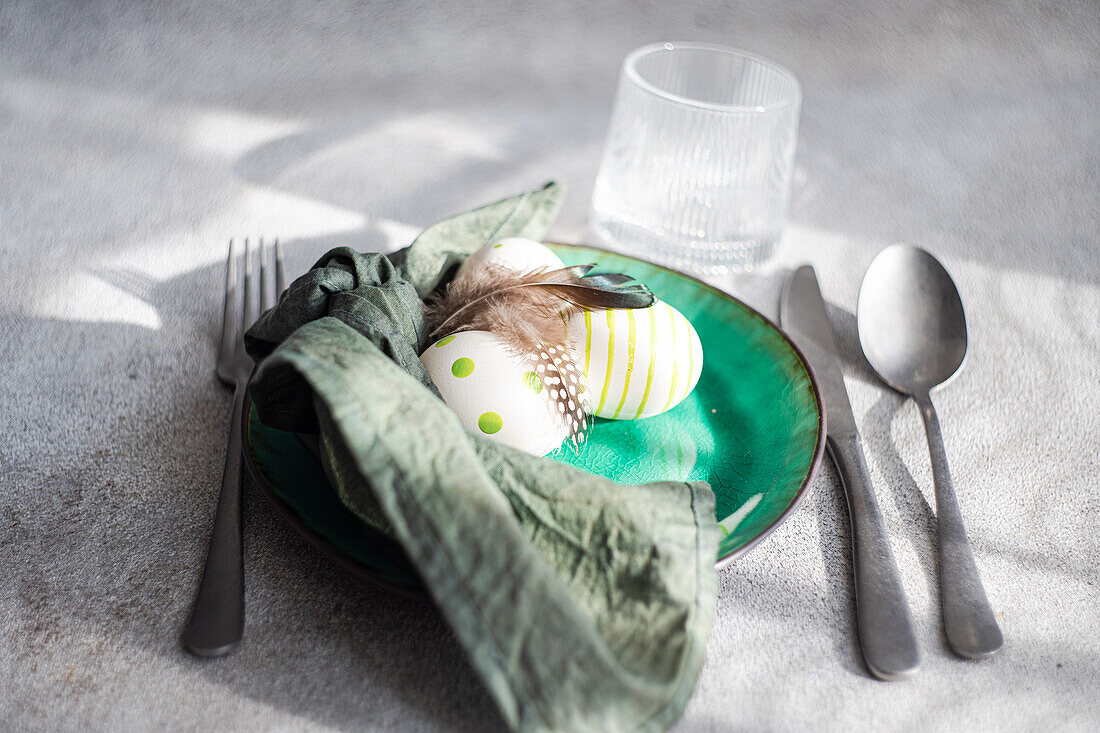 Closeup of Easter table setting, showcasing a vibrant green ceramic plate with two decorative Easter eggs adorned with white and green patterns and delicate feathers, placed on gray surface between napkin and cutlery and glass of water