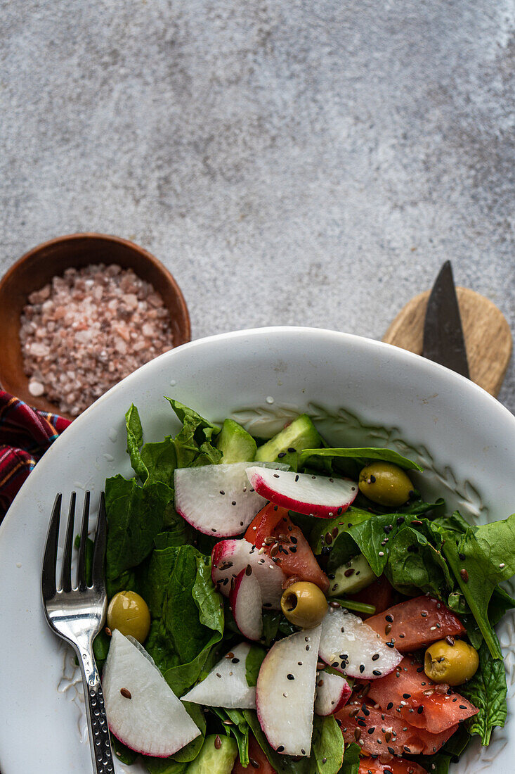 A vegetable salad made with fresh organic spinach leaves, sliced tomatoes, radishes, and olives presented in a white bowl beside a plaid cloth, wooden cutting board, and a bowl of pink salt