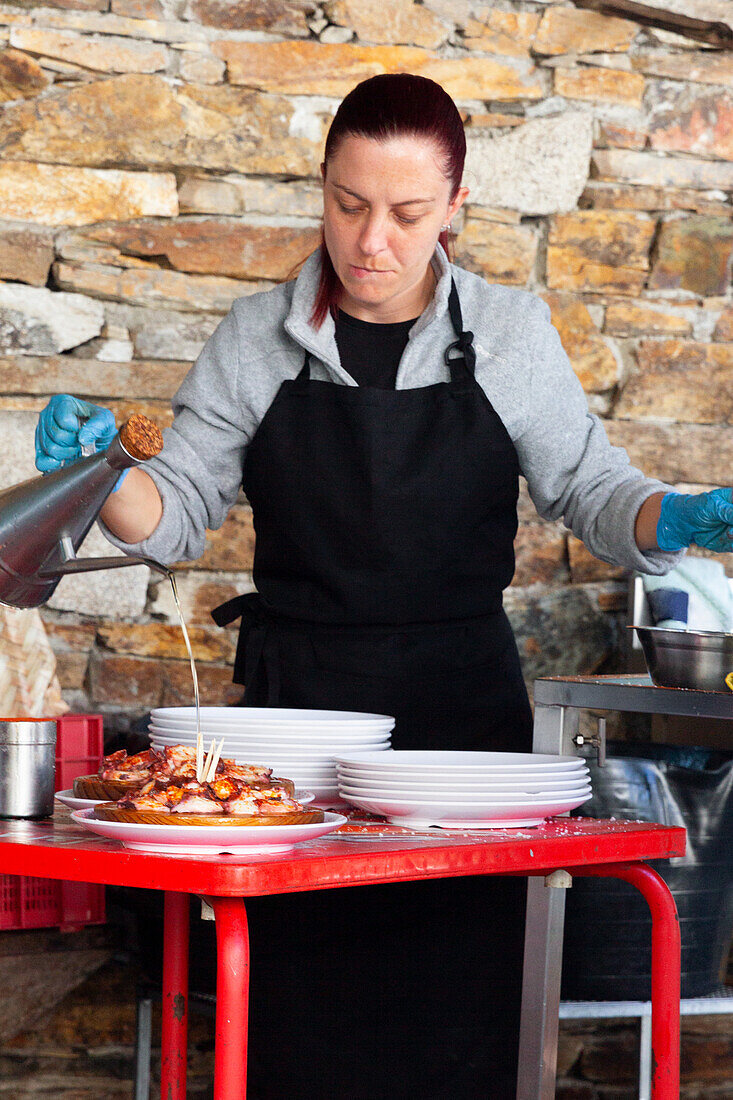 Konzentrierter Koch mit schwarzer Schürze und Handschuhen träufelt sorgfältig Olivenöl über einen Teller mit köstlich gegartem Oktopus vor einem rustikalen Steinmauer-Hintergrund