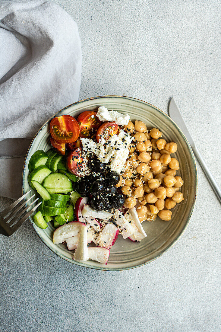 Top view of bowl with healthy salad and vegetables with chickpea sesame seeds and olives with cutlery and fabric placed on bright gray table