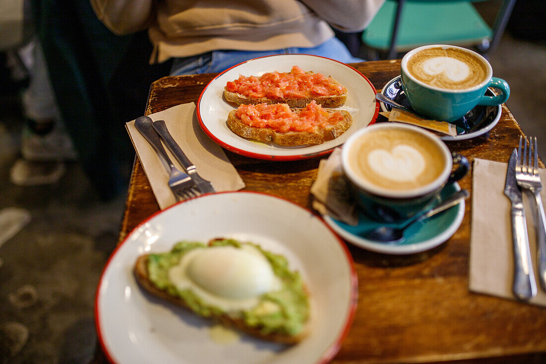 Delicious toast with poached egg and smashed avocado on ceramic plate near cups of cappuccino in cafe