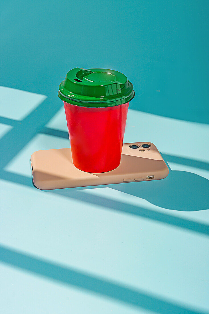 Minimalistischer roter Plastikbecher mit Kaffee und grünem Deckel auf einem Tisch über einem Mobiltelefon vor blauem Hintergrund