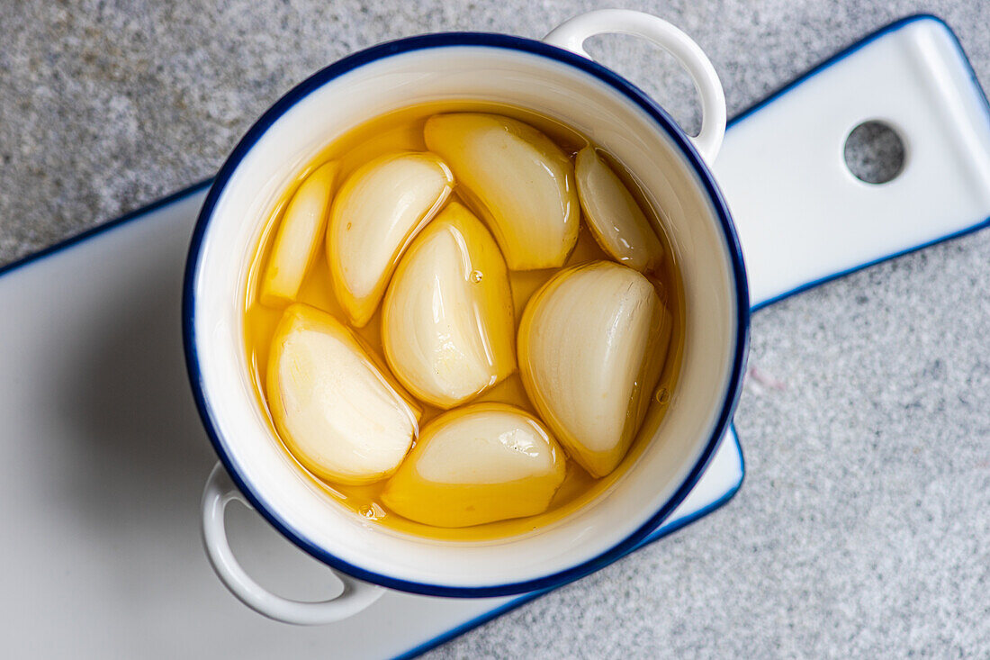 Top view of white ceramic pot filled with peeled and baked garlic cloves drenched in golden oil, positioned on a gray surface with a blue-trimmed handle visible