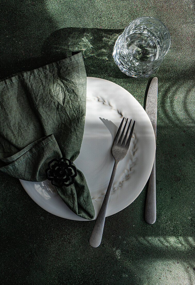 An artistic table setting featuring a white plate with a decorative napkin, utensils, and a clear glass on a table casting soft shadows.