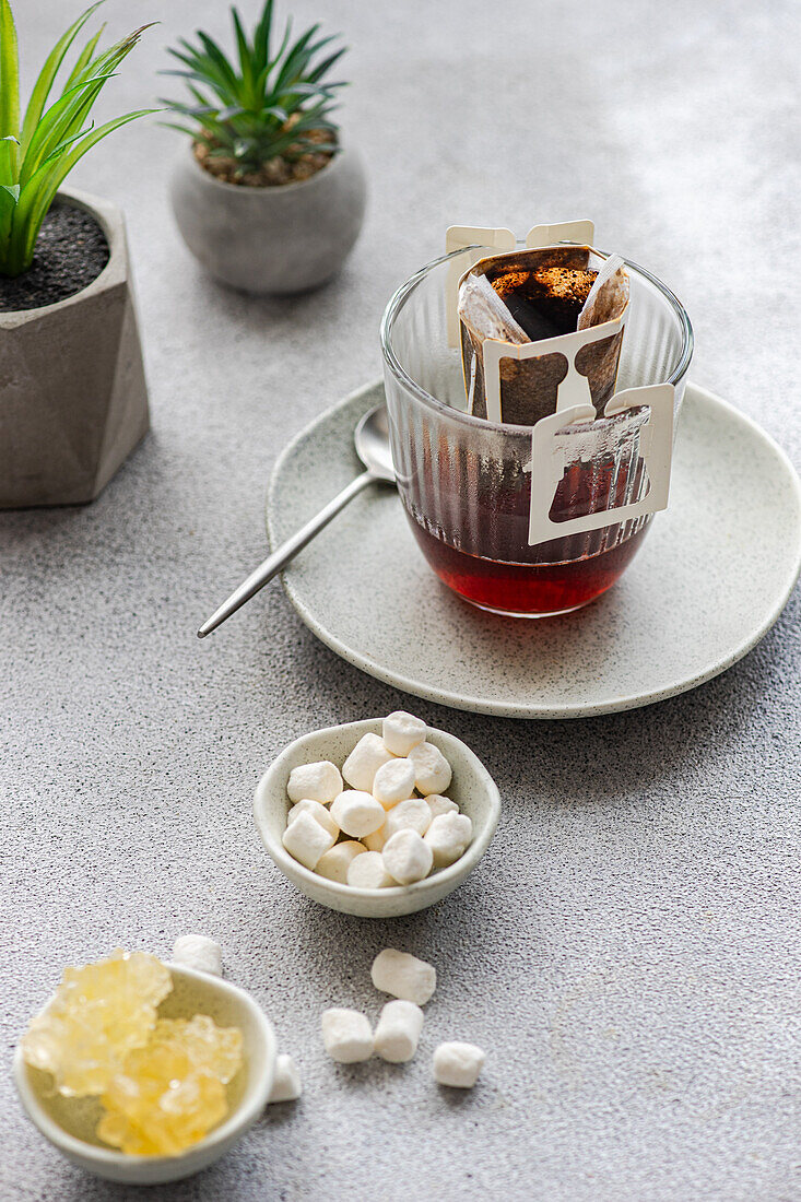 A fresh cup of drip coffee brewing in a clear glass with trendy grey backdrop, accompanied by sweet marshmallows and chic decor plants.