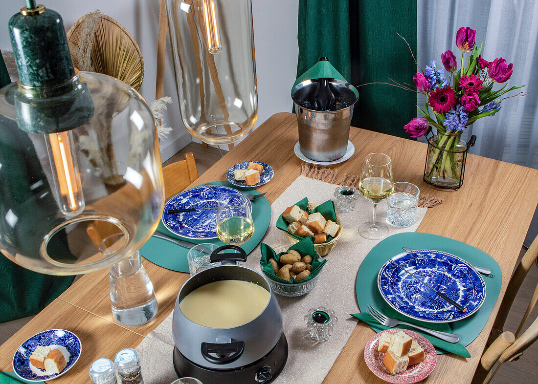 An intimate dining setting features a cheese fondue pot, wine glasses, ornate blue plates, and fresh flowers, offering a cozy ambiance for a gourmet experience.