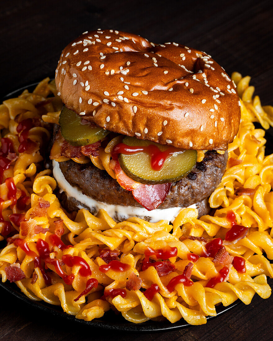 Hoher Winkel eines Hamburgers auf einem Teller mit Makkaroni, Ketchup und Käse vor dunklem Hintergrund