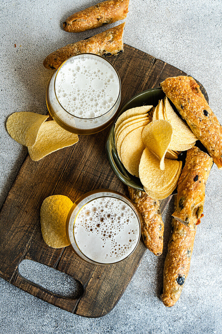 Top view of tasty pieces of crisp potato chips in bowl with bread sticks and glasses of beer on wooden board placed over table in daylight