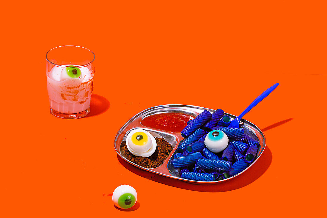 Horror-Mittagessen mit bunten Nudeln auf einem Tablett vor orangefarbenem Hintergrund neben einem Glas mit weißer Flüssigkeit und einem Auge