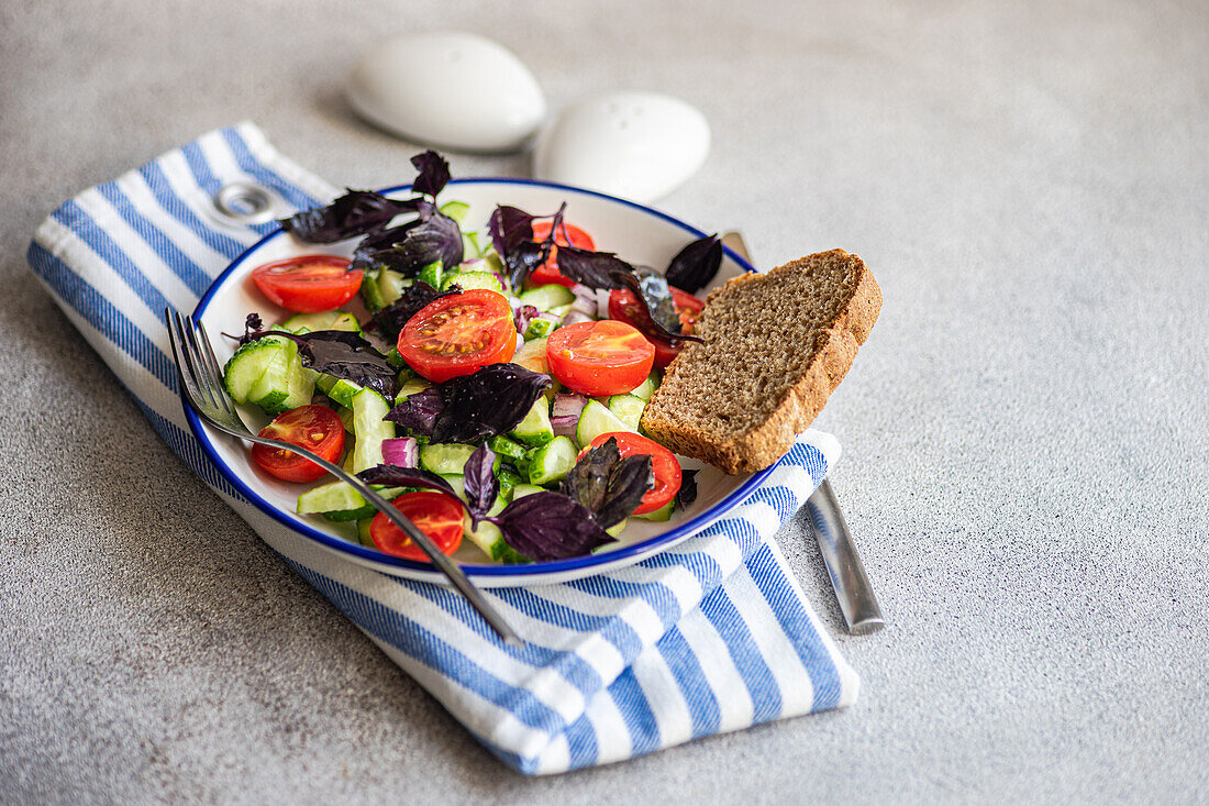 High angle of Seasonal vegetable salad with organic cucumber, tomato, onion and red basil leaves served on plate placed on striped napkin against gray background