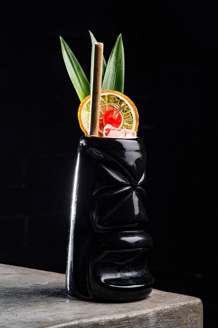 Refreshing alcoholic cocktail garnished with green leaves lemon slice and cherry served in black Tiki mug placed on stone table