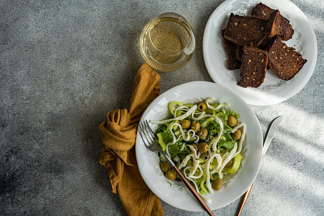 Top view of Vegetable salad with green vegetables like lettuce, cucumber, olives, green bell pepper with homemade cheese and sesame seeds placed near plate of bread, glass of wine and cutlery against gray background