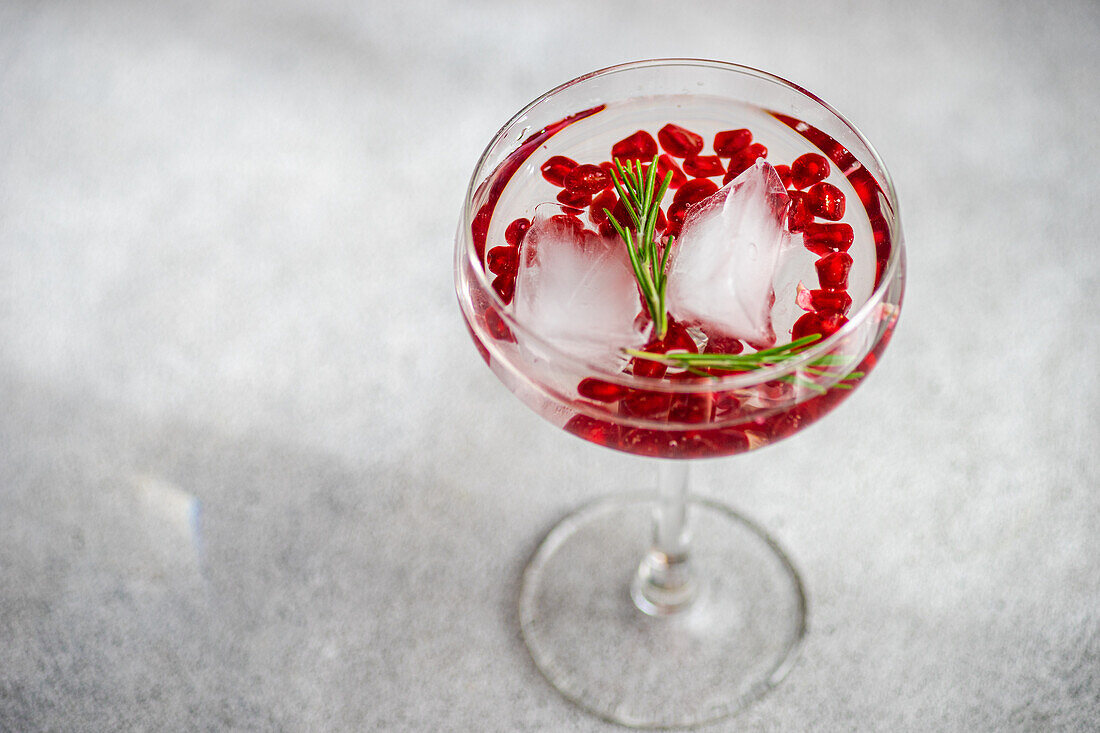 Gin-Tonic-Cocktail von oben mit Granatapfelkernen und Rosmarin in einem breitrandigen Glas auf grauem Hintergrund