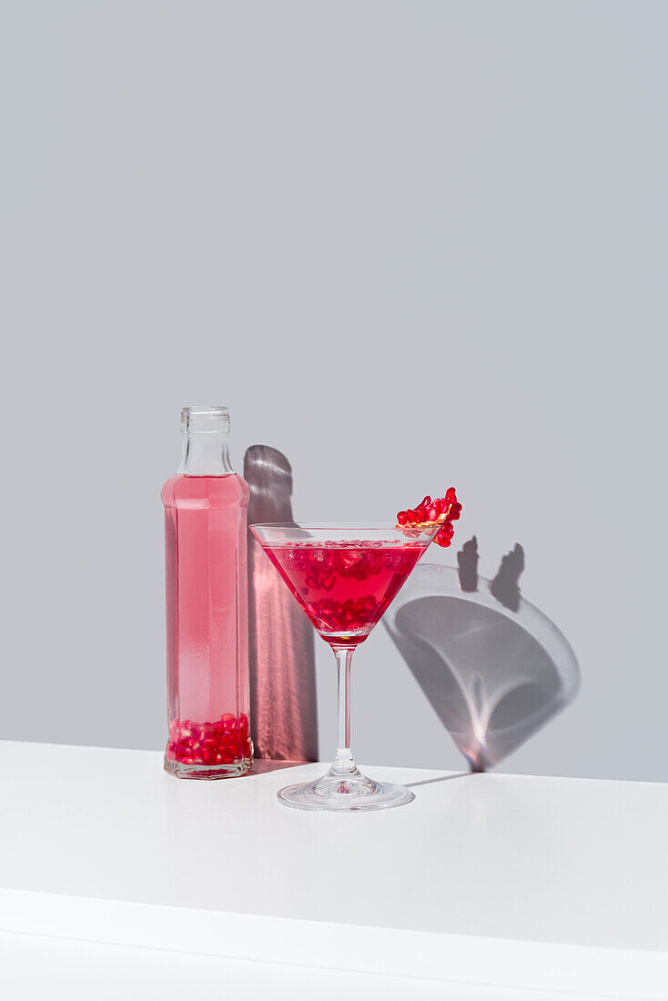 Glass filled with red pomegranate cocktail and adorned with pomegranate seeds, stands beside a transparent bottle of the same vibrant drink, both casting soft shadows on a calm gray background