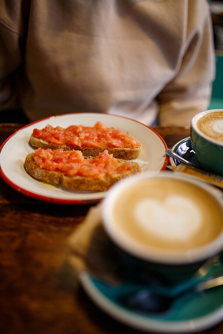 Köstliche, appetitliche Toasts mit Tomaten auf einem Teller neben Tassen mit aromatischem Cappuccino auf einem Holztisch in einer Cafeteria