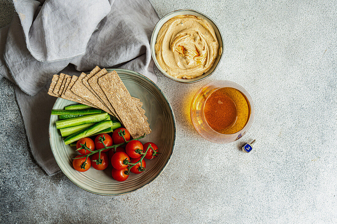 Draufsicht auf einen gesunden, pflanzlichen Teller mit Hummus und Gemüse, serviert in Schalen neben einer Serviette und einem Glas Schnaps vor grauem Hintergrund