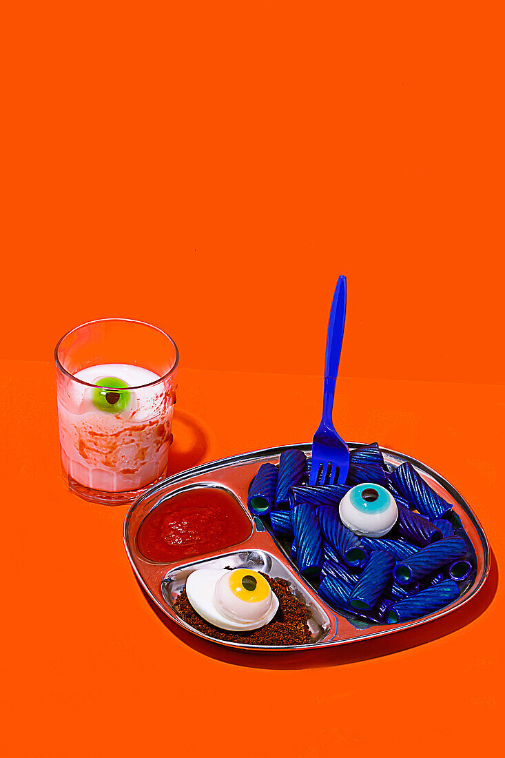 Hoher Winkel des Horror-Mittagessens mit bunten Nudeln auf einem Tablett vor orangefarbenem Hintergrund neben einem Glas mit weißer Flüssigkeit und einem Auge