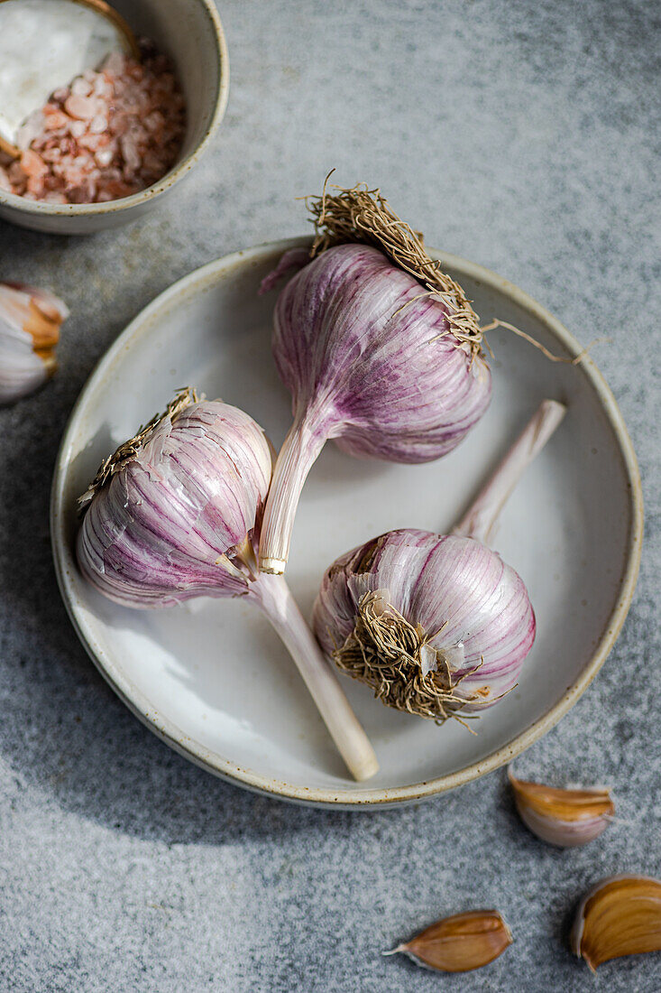 Top view of shot of raw garlic bulbs with purple streaks set against a grey background alongside a dish with coarse salt