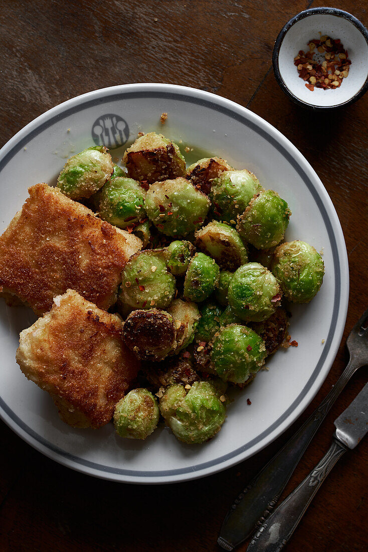 A rustic meal consisting of golden brown, pan-fried brussels sprouts and crispy fish fillets, served on a ceramic plate with a side of chili flakes