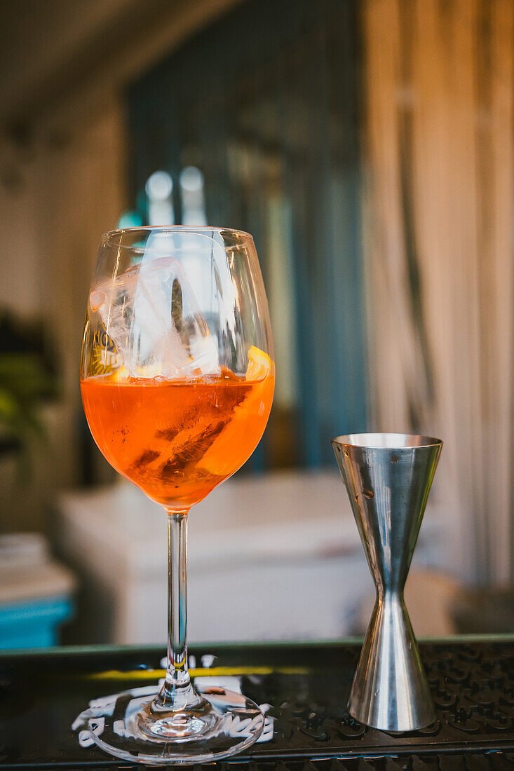 Orange cocktail in a wine glass on a bar, with a jigger and coaster in soft focus background