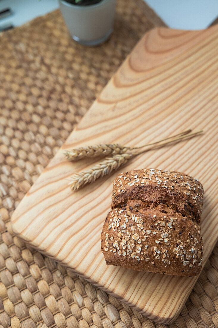 Ein Laib Saatenbrot neben einer Weizenähre, auf einem Holzbrett über einer gewebten Matte, mit natürlichen Zutaten