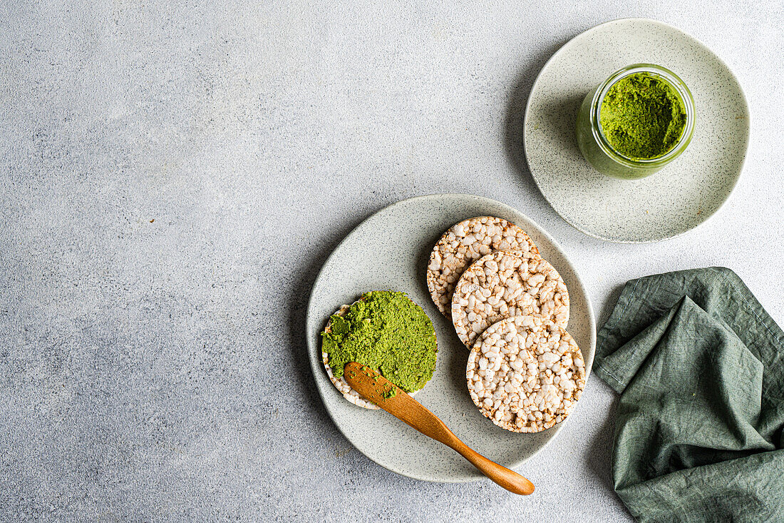 Draufsicht auf Reisbrot auf einem Teller mit einer leuchtend grünen Spinat-Pesto-Pasta-Soße in einem Glas, vor grauem Hintergrund neben Serviette und Salzstreuer