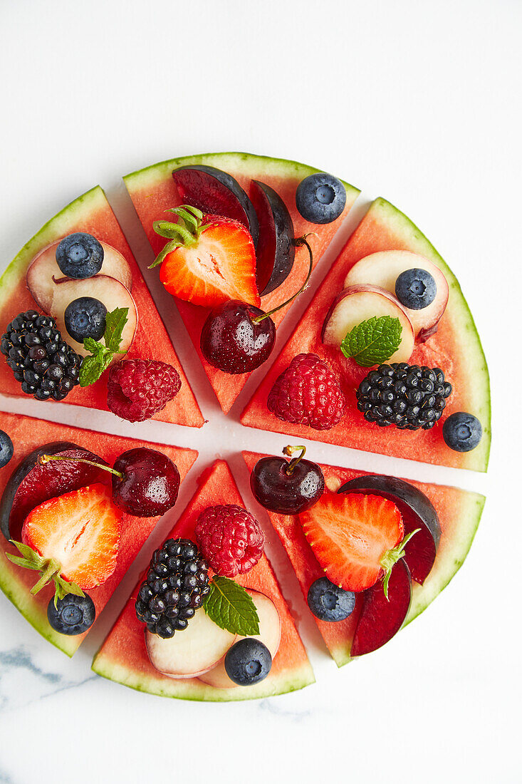 Top view of ingredients consisting of strawberry apple raspberry grapes blackberry plum blueberry peach placed on round cut slices on white surface while preparing watermelon pizza
