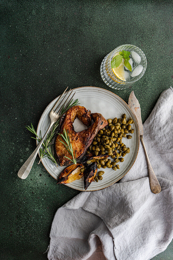 Top view of sunlit healthy lunch setup featuring BBQ salmon steak seasoned with spices alongside roasted lemon and a serving of fermented capers