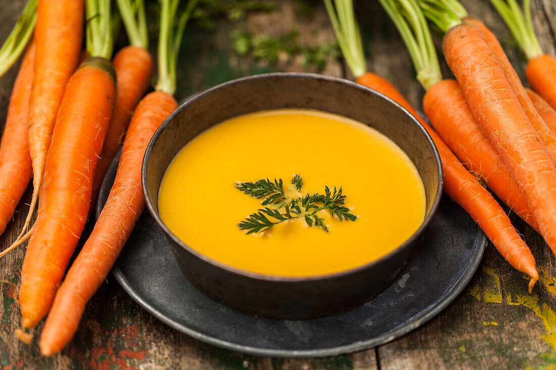 Fresh carrot soup with parsley in a black bowl, bunch of carrots on the side, wooden surface.